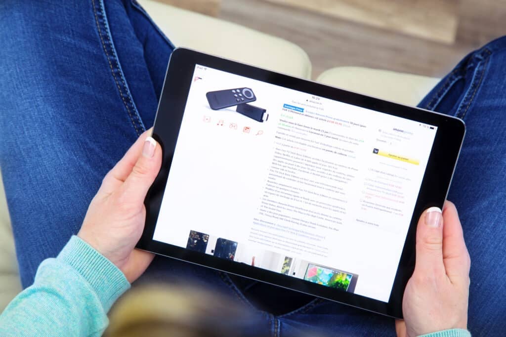 A woman sits cross-legged on her couch, using a touchpad to browse products on the Amazon website.