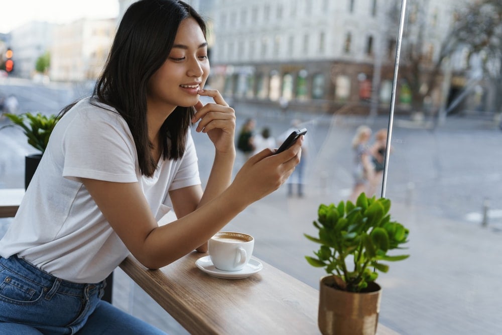 A young, attractive Asian girl sits by the window in a trendy cafe with an espresso cup beside her, checking her phone.