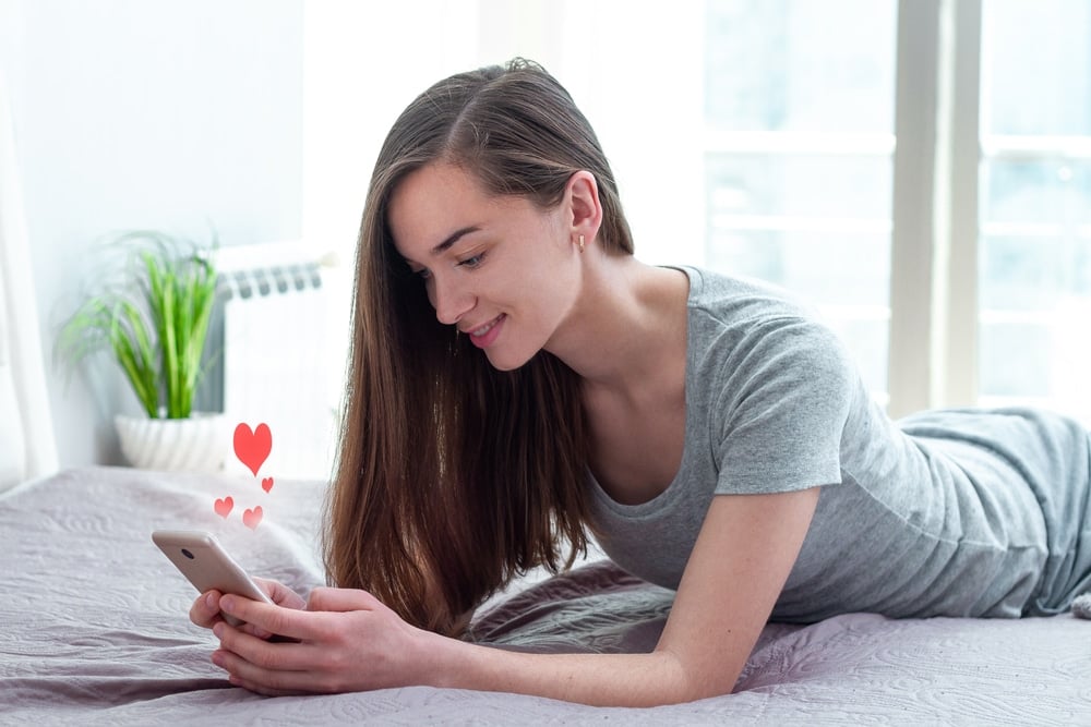 A young, attractive brunette lady lies stomach-down on her bed while checking her phone, with hearts emanating from it.