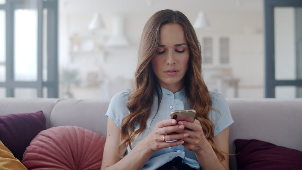 An attractive woman in a short-sleeve shirt sits on a sofa, checking her smartphone.