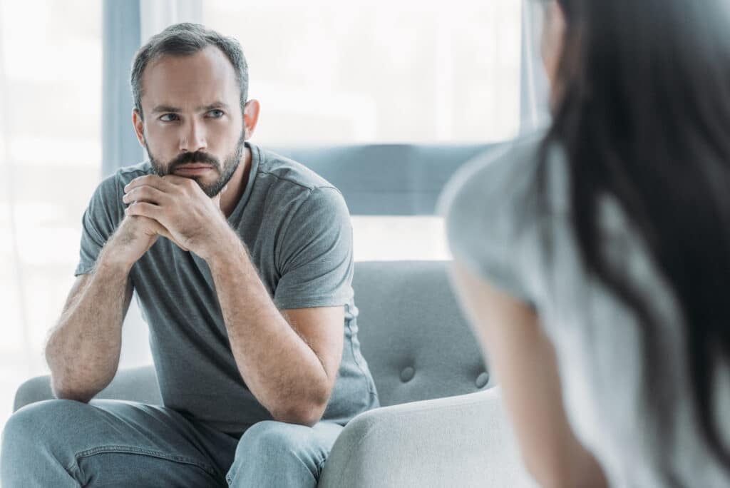 A man sits on a sofa chair, leaning forward with his hands to his chin, deep in thought as he looks over to a lady.
