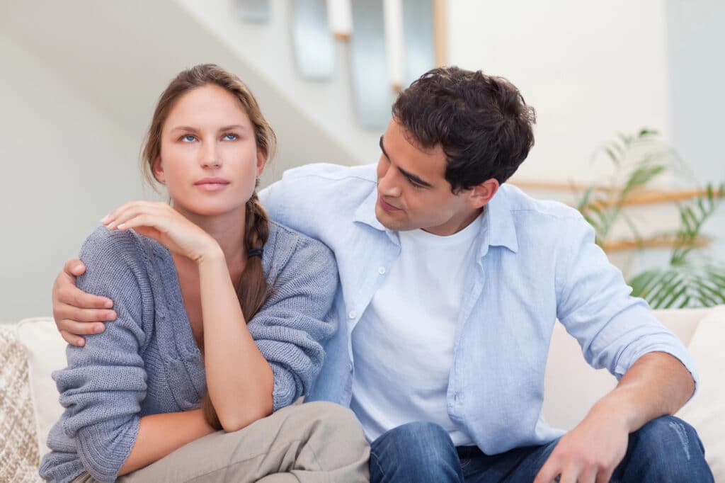 A young couple sits on a sofa together. The man puts his arm around the woman, trying to cheer her up, while she looks slightly annoyed.