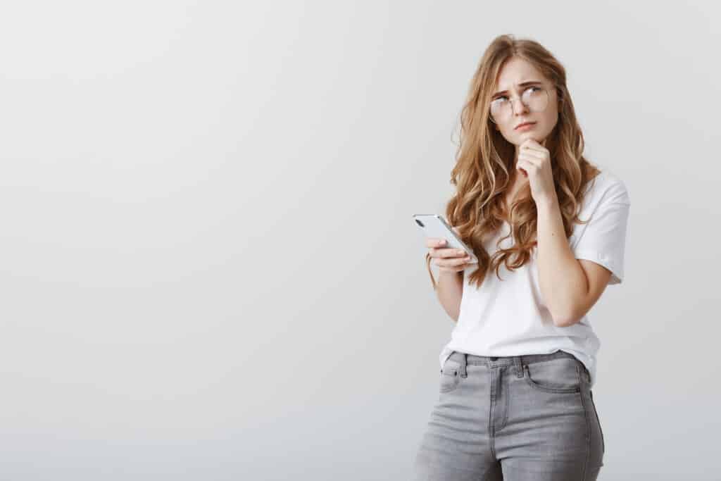 A cute, ordinary European girl in glasses is holding a smartphone, touching her chin, and frowning while looking up thoughtfully.