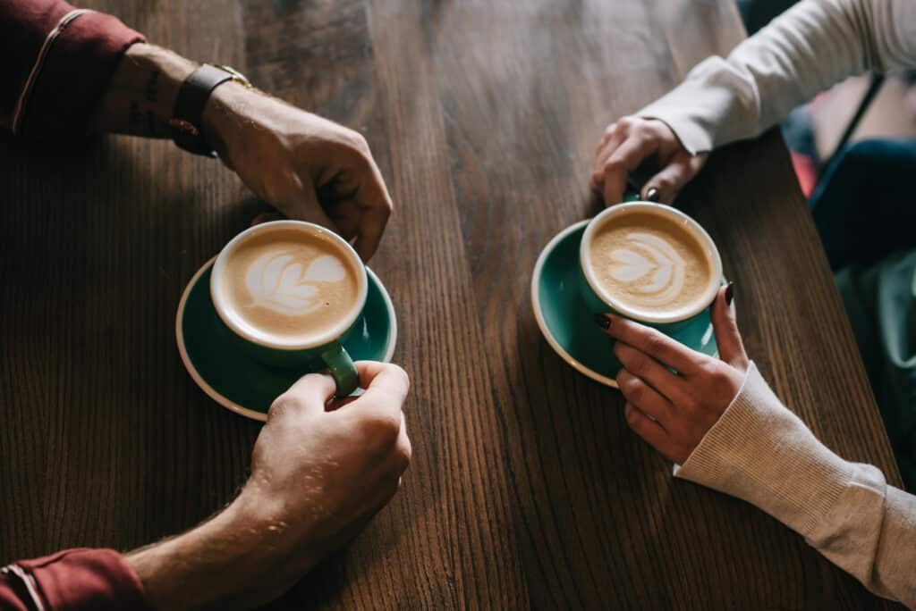 A close-up of a couple's hands sitting across from each other, drinking coffee with a heart-shaped foam.