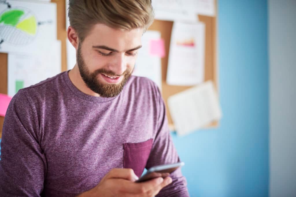 A man with a beard, wearing a purple shirt, typing on his smartphone in his office.