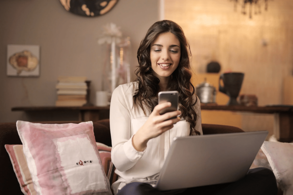 A young girl smiles as she checks her phone, with her laptop on her lap while sitting on a sofa.