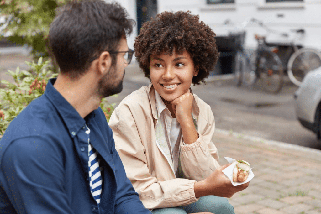 A mixed-race couple looks happily at each other as they sit outside, while the lady holds a hotdog.