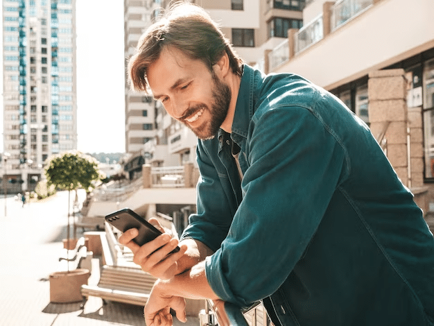 A handsome, bearded young man smiles as he leans on a rail outside, checking his phone.