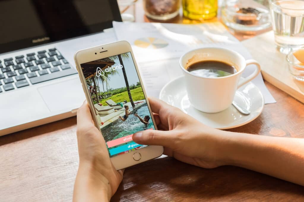 "Airbnb books: Someone is scrolling on their Airbnb app on their phone in front of their laptop with a coffee at a cafe.