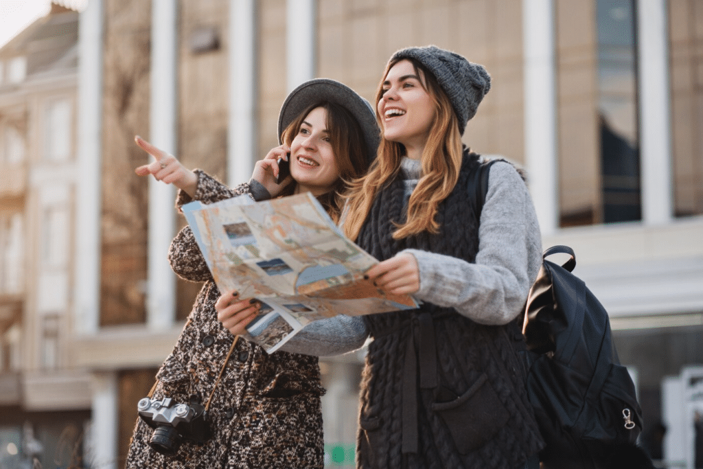 Two young female tourists are smiling and looking in the distance, with one holding a map and the other pointing at something.