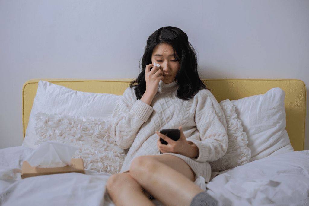 A young Asian woman lies on her bed, wiping away her tears with a tissue as she looks at her phone.