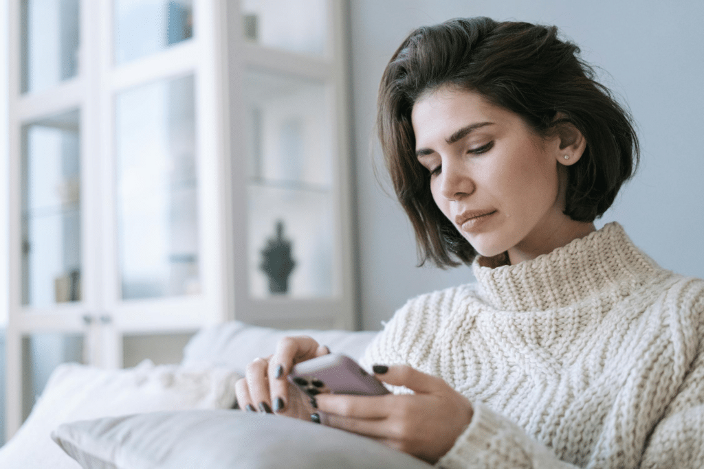 A brunette-haired woman in a white sweater looks sad as she stares down at her phone on the sofa.