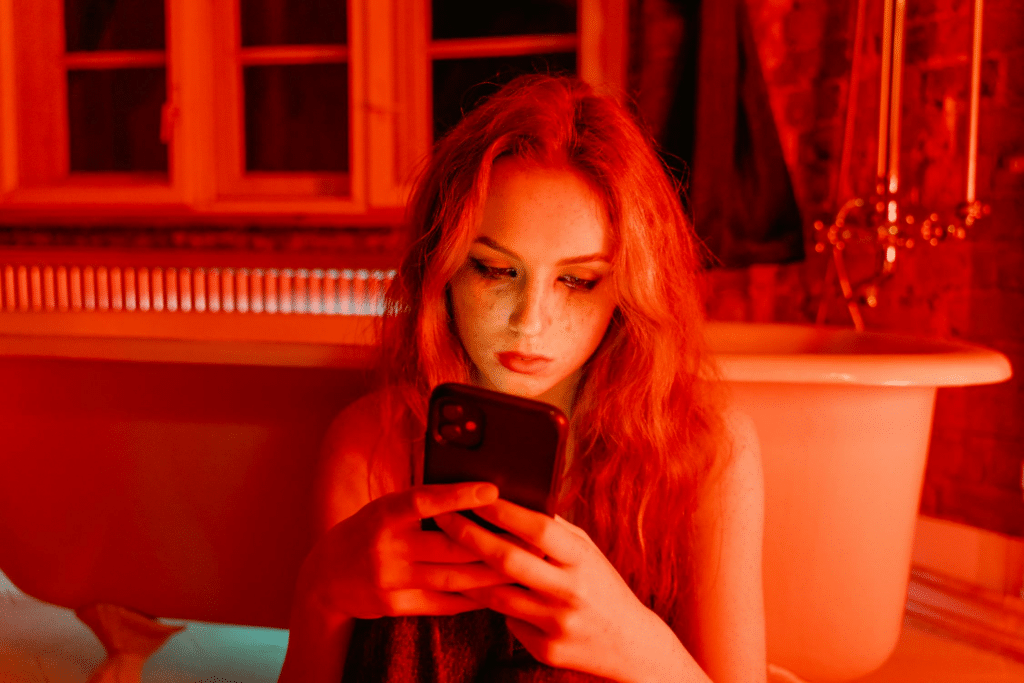 A young girl sits in the bathroom in front of her tub, staring at her smartphone while the room is illuminated in red light.