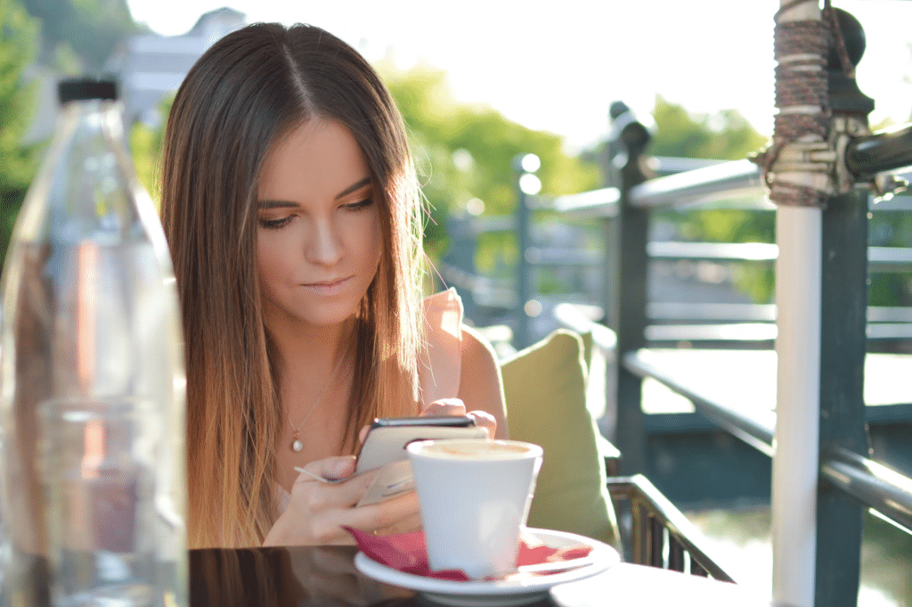 A young woman sitting in outside restaurant with an espresso and water typing on her phone.
