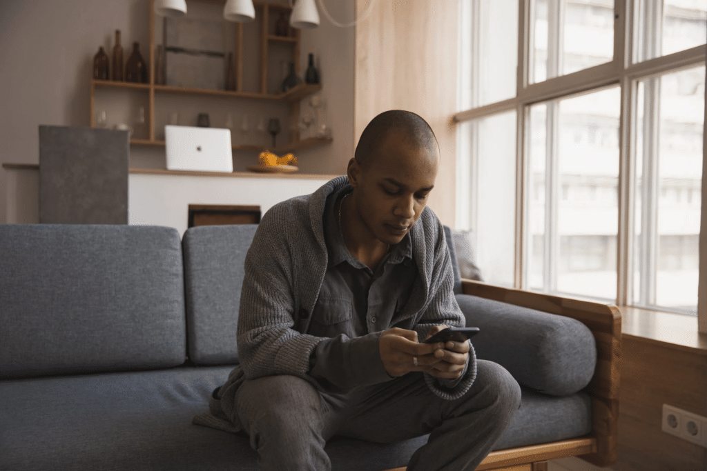 Man in a sweater sitting on his sofa by the window, checking his phone.