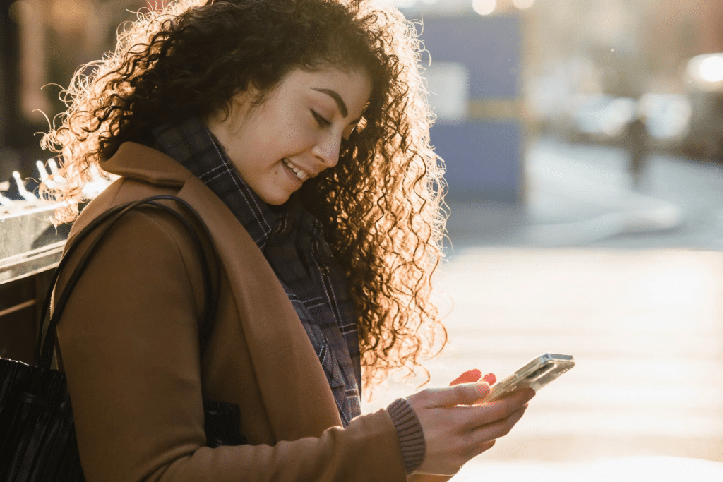 A young woman with curly hair, wearing a stylish jacket and scarf, smiles as she checks her phone outside.