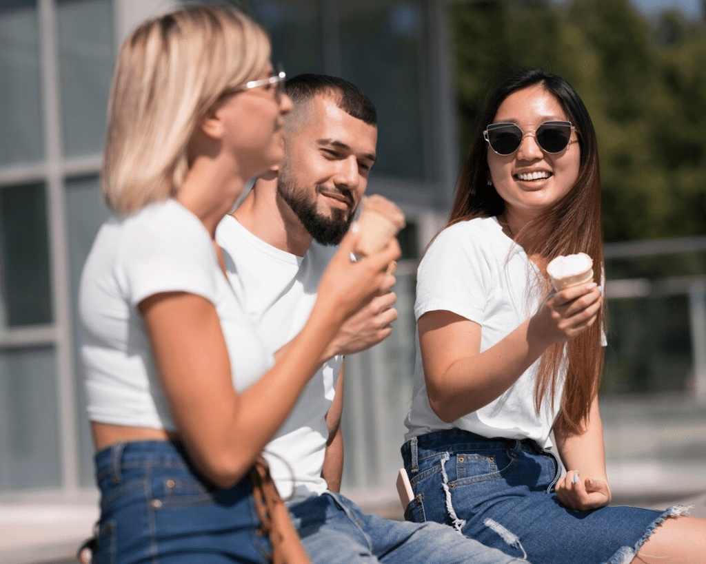 A guy and two female friends hang out outside on a sunny day, enjoying ice cream.