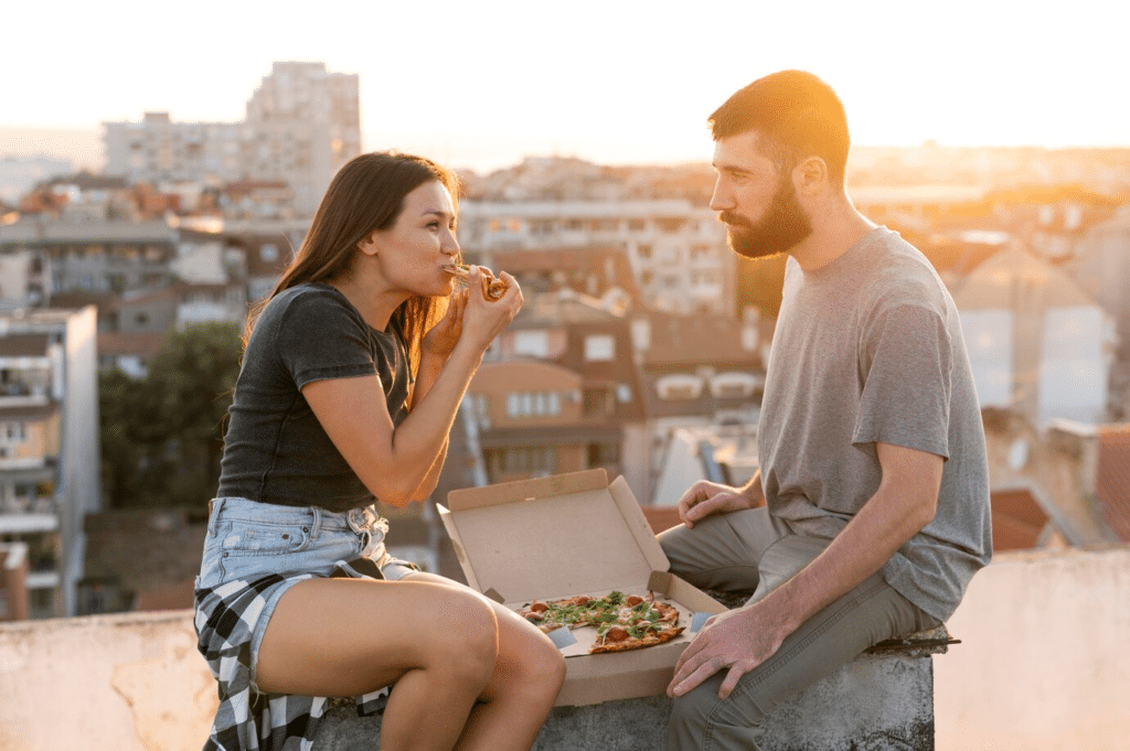 A young couple sit on a rooftop enjoying a box of pizza with the sunset and buildings behind them.