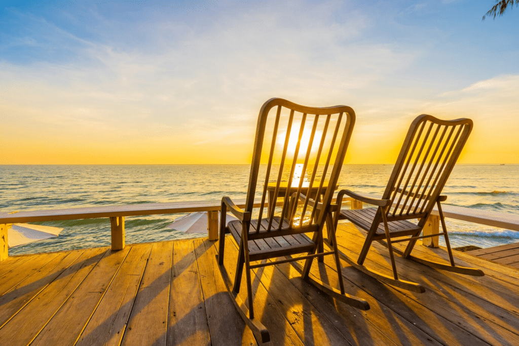 Two wooden recliners face the water on a deck, with the sunset visible in the distance.