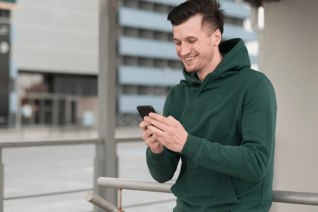 A young man in a green hoodie smiling as he texts on his phone outside.