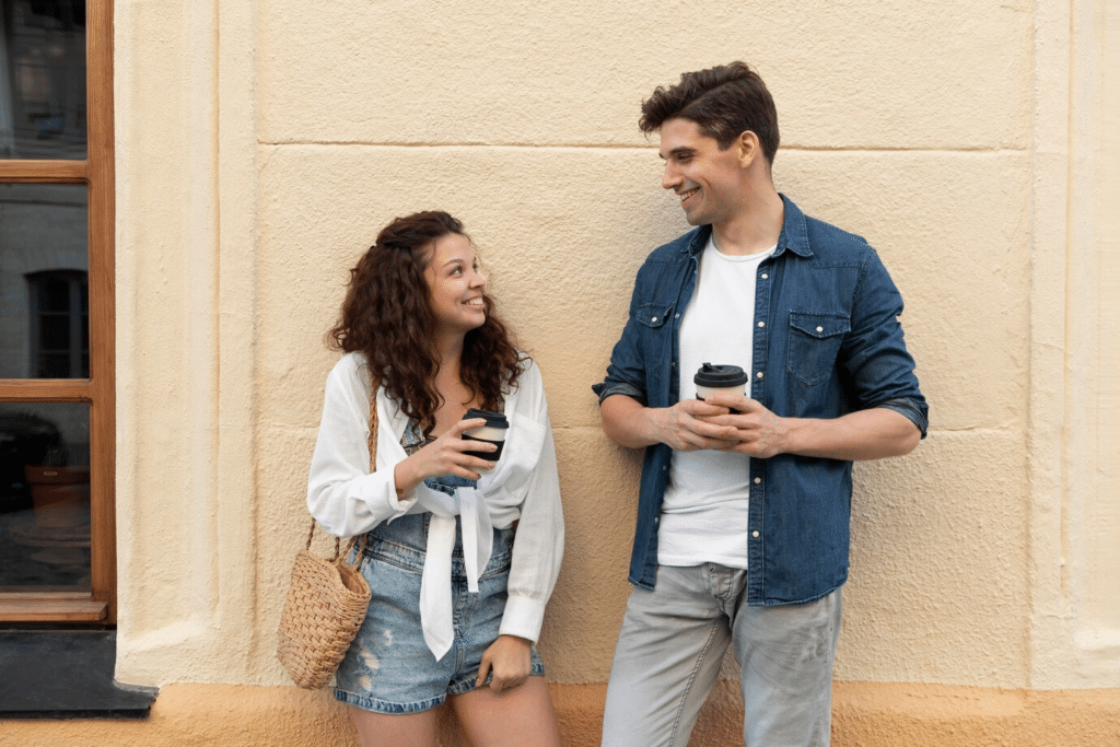 A young man and woman chatting and smiling as they enjoy coffee outside in front of a building.