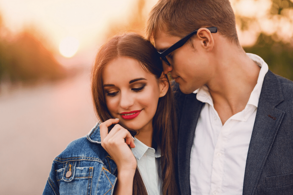 A young couple stands outside. The man, wearing glasses, is seen smelling the woman's hair as she looks away shyly, smiling.