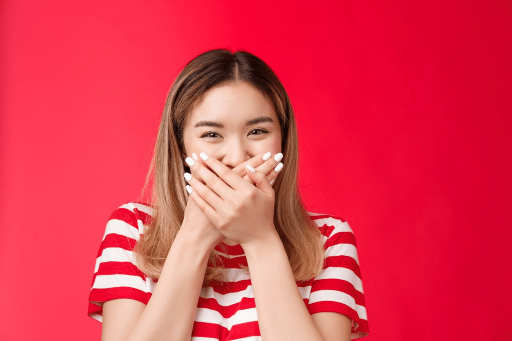 A young Asian girl covers her mouth as she giggles against a red background.