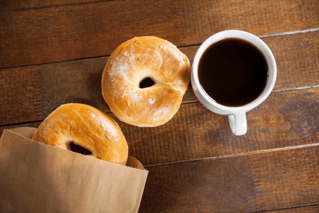 A mug of black coffee sits beside two bagels on a wooden table.