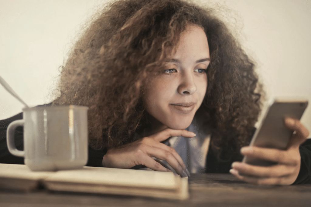 A young lady with curly hair leans on her desk, checking her phone with her mug and notebook in front of her.