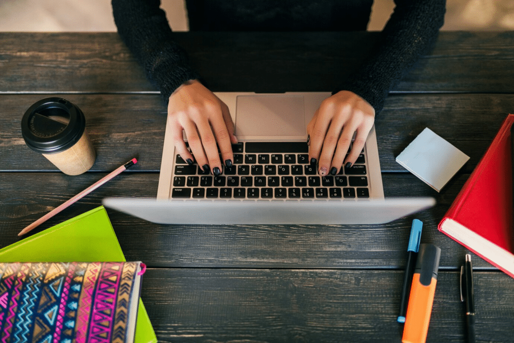 Hands of a female typing on her laptop, with a coffee cup, notebooks, pens, and markers on a wooden desk."